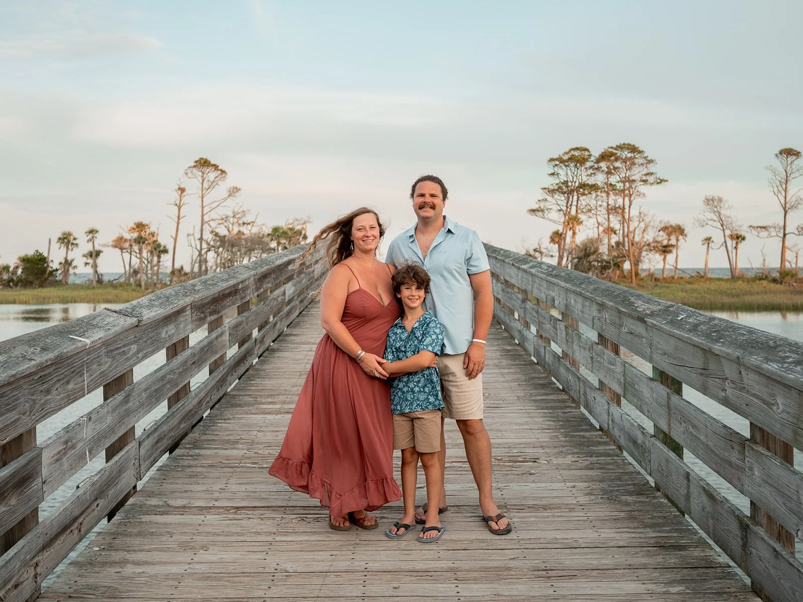 Family walking along a wooden pier with marsh grasses on either side, holding hands and smiling.