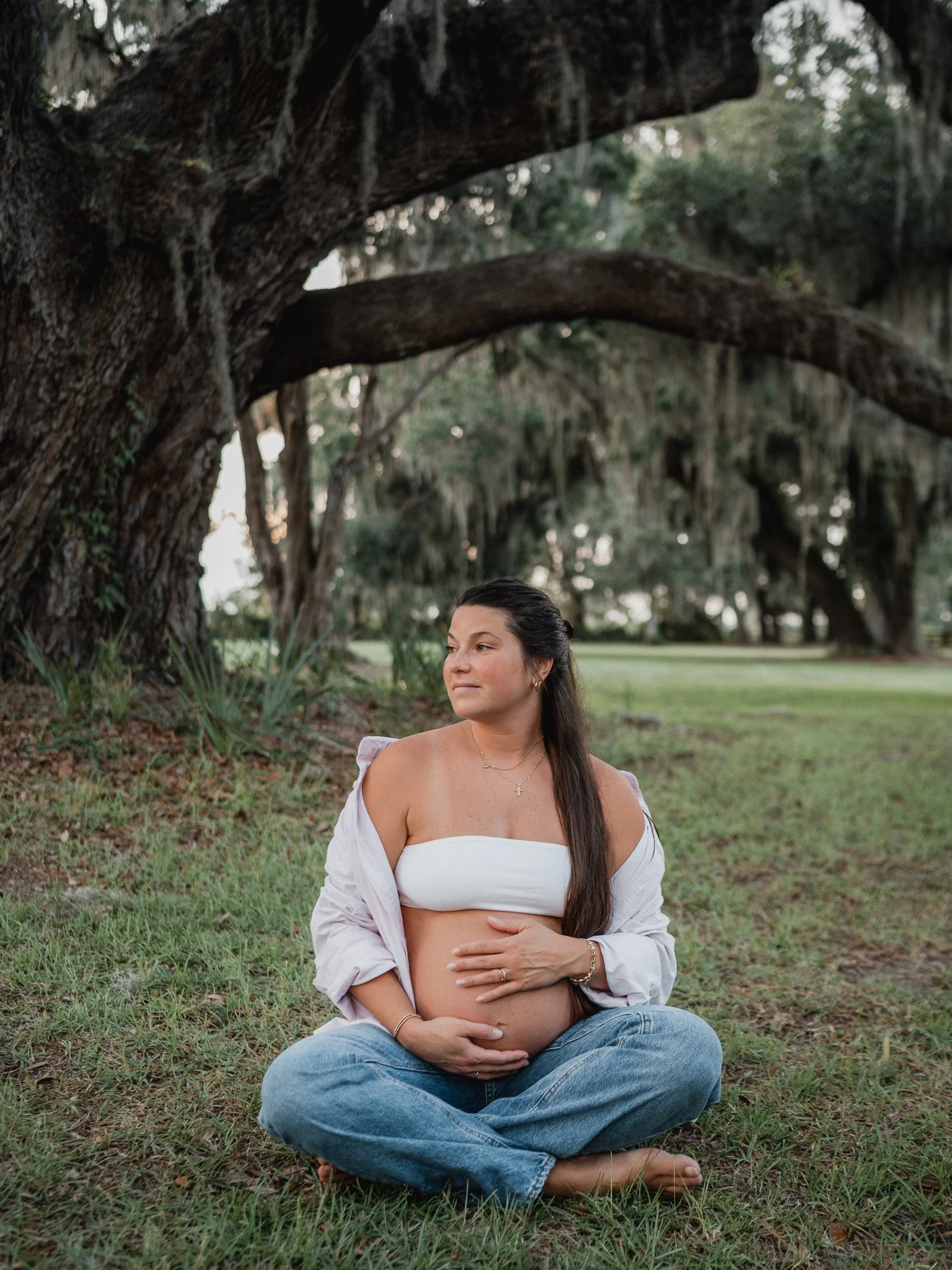 Expectant woman seated under a Lowcountry oak tree, barefoot and holding her pregnant belly with both hands.