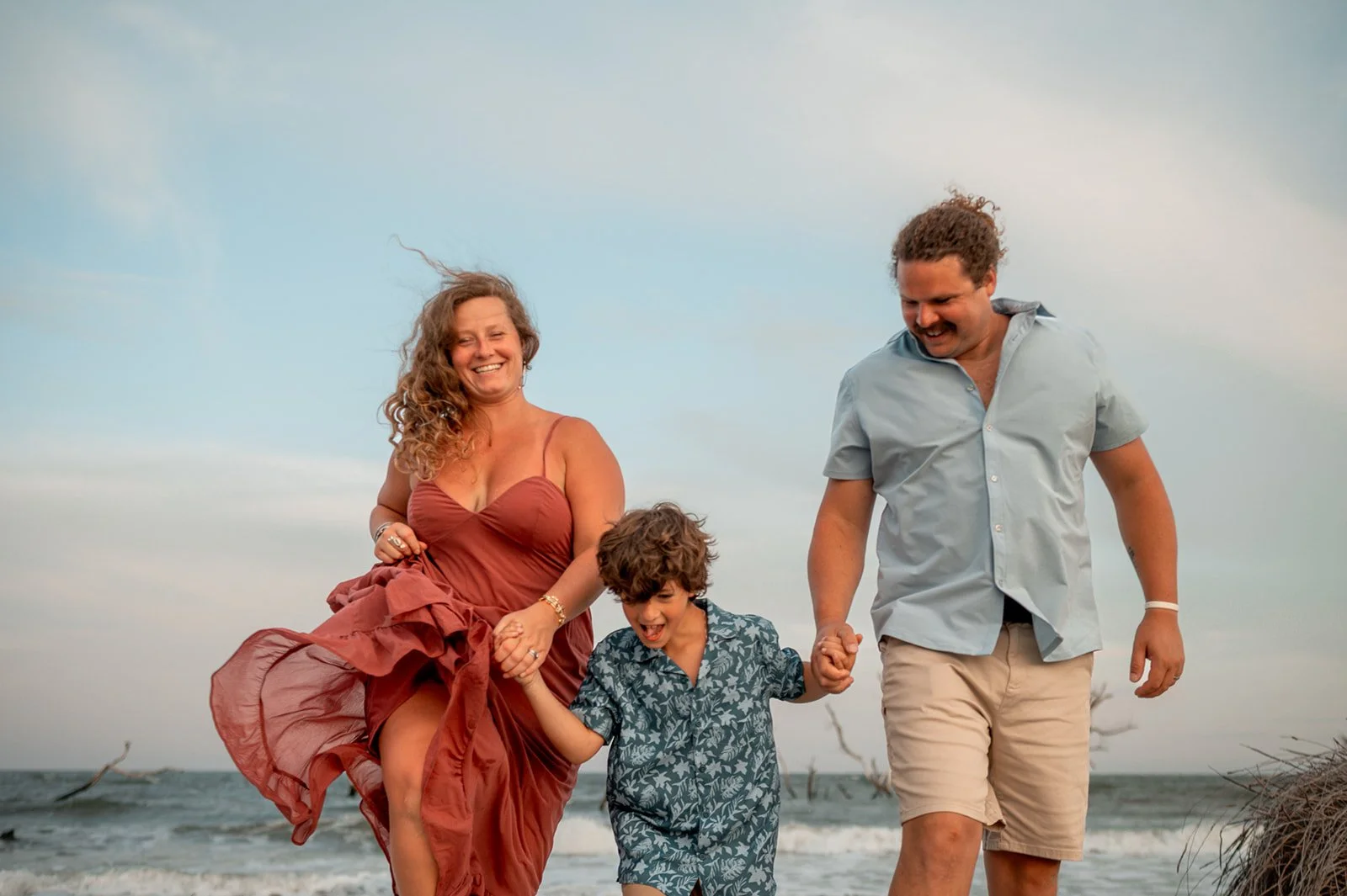 Family walking on a beach boardwalk with dunes on either side; young child skipping ahead.