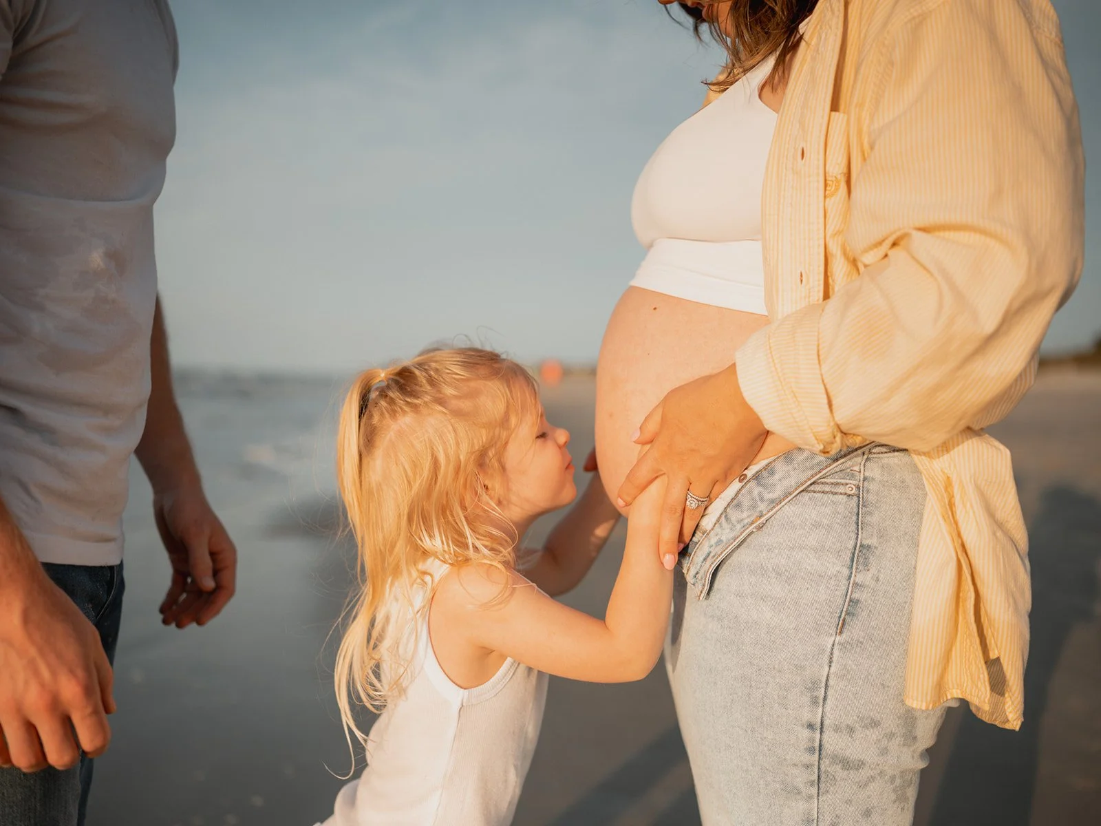 Young girl hugging her mother’s pregnant belly at sunset in a grassy field, bathed in golden light.