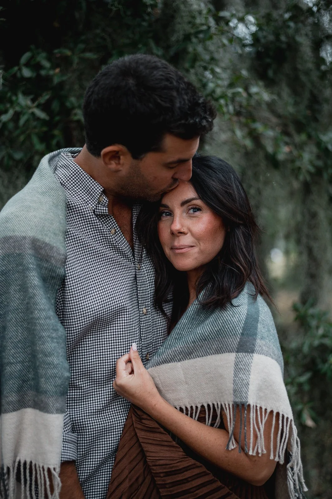 Couple wrapped in a plaid blanket, man kissing his fiancé on the forehead with oak trees in the background.  