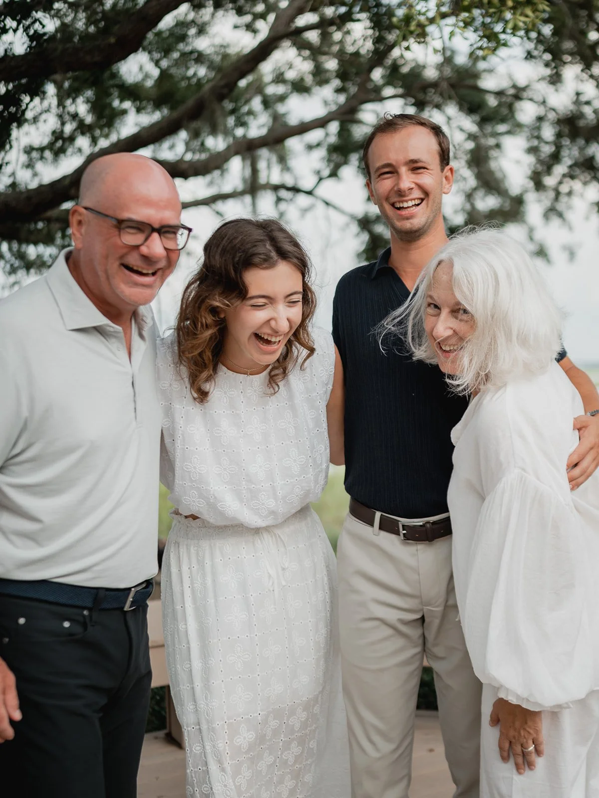 Extended family group hugging and laughing together outdoors, trees and Spanish moss behind them.