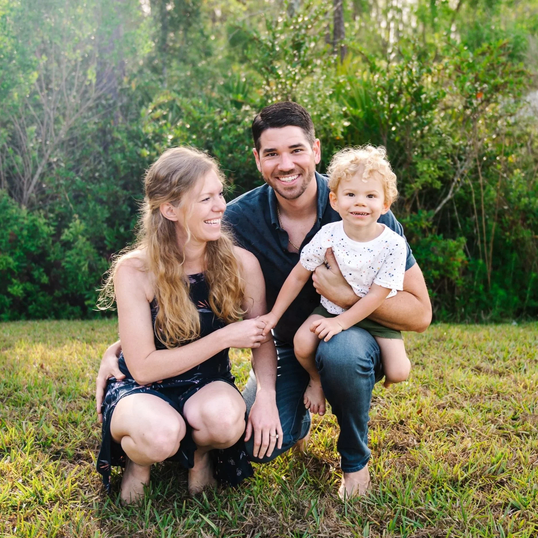 Family sitting together on a grassy field, parents embracing their young child during a Lowcountry golden hour.