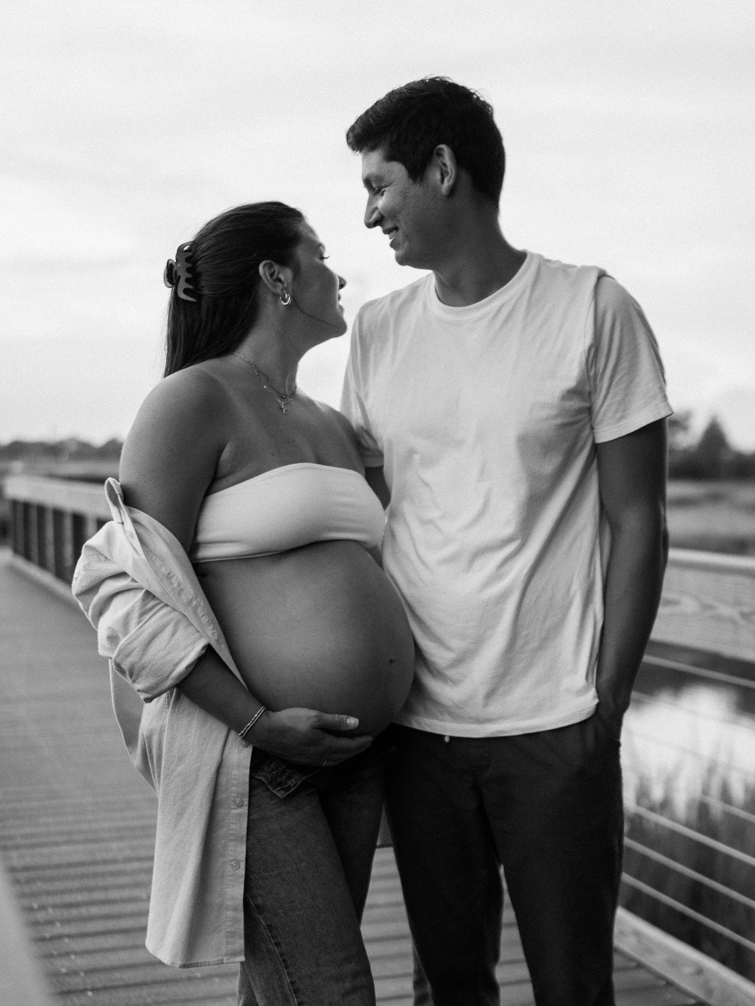 Expectant couple standing on a wooden boardwalk by the marsh; the father cradles the mother’s baby bump.