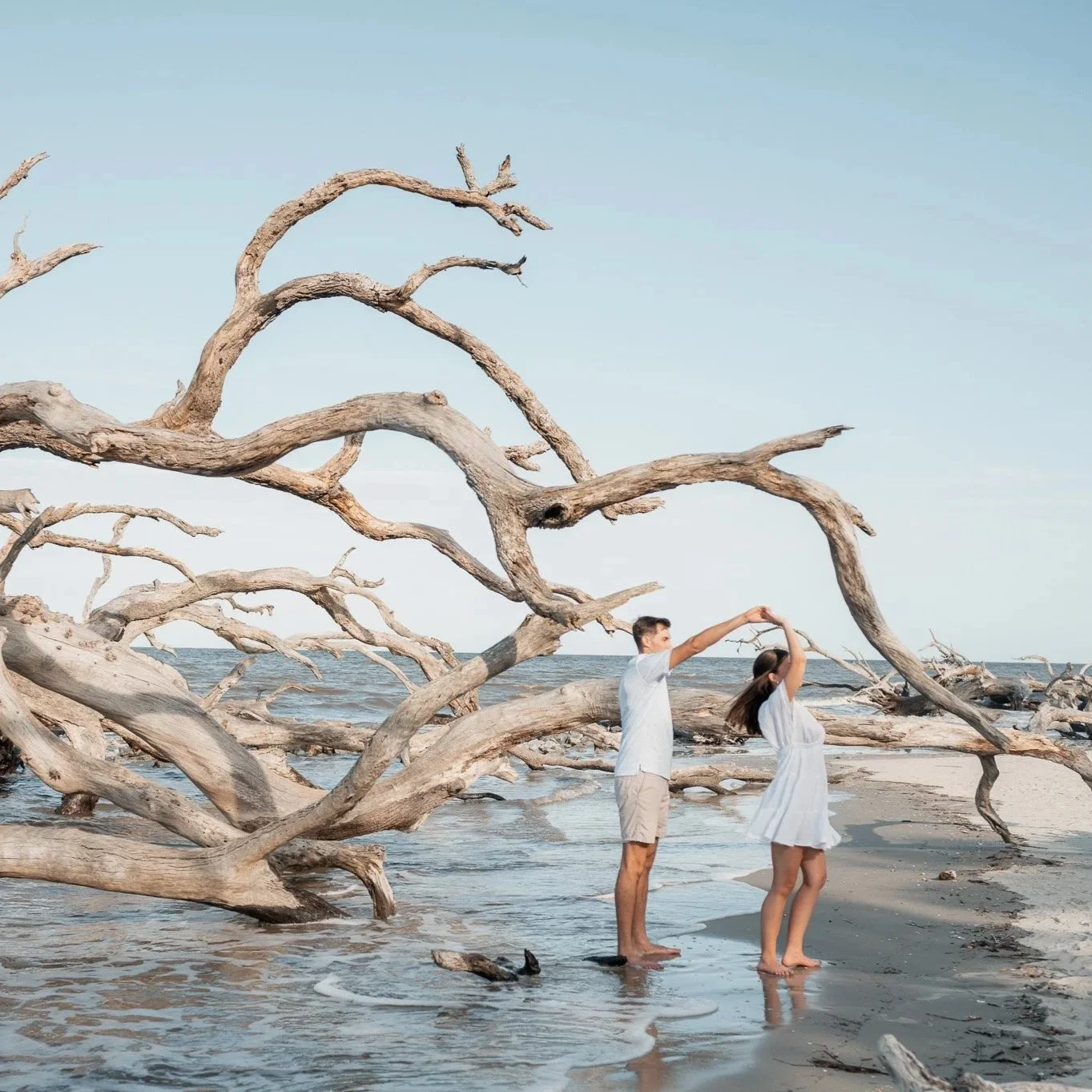 Couple standing hand‑in‑hand on Hunting Island driftwood beach at dusk, soft pink sky and ocean waves in the background.