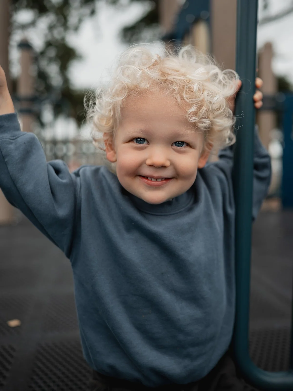 Close‑up of a toddler with curly blond hair laughing while sitting on the beach.