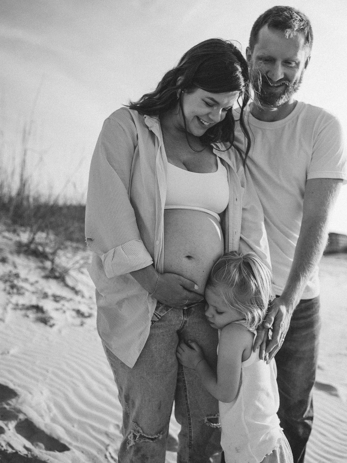 Black‑and‑white family portrait on a beach: parents embrace while their toddler hugs the mother’s pregnant belly.