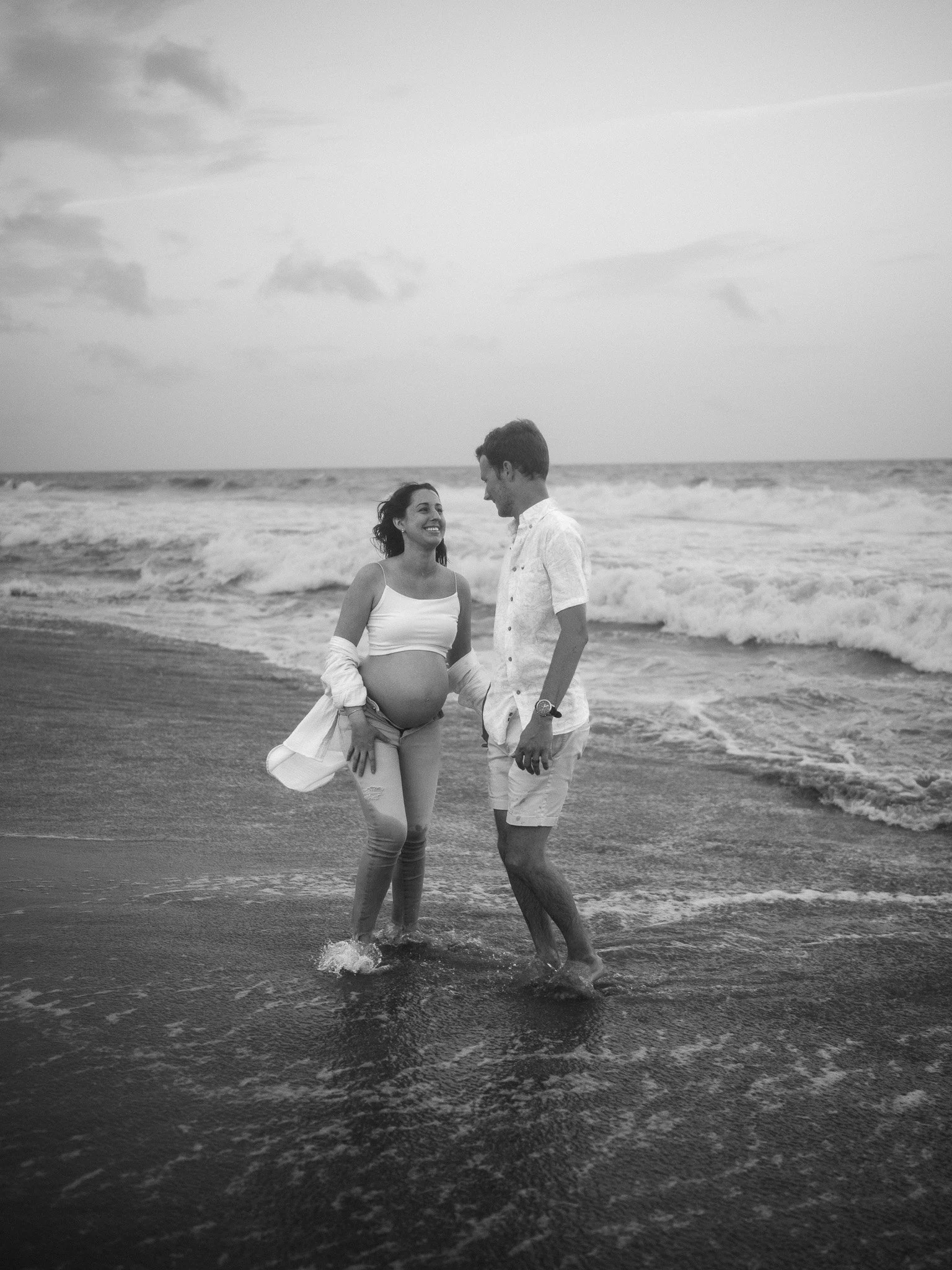 Black‑and‑white photo of an expectant couple walking along the shoreline, the father’s hand resting on the mother’s baby bump.