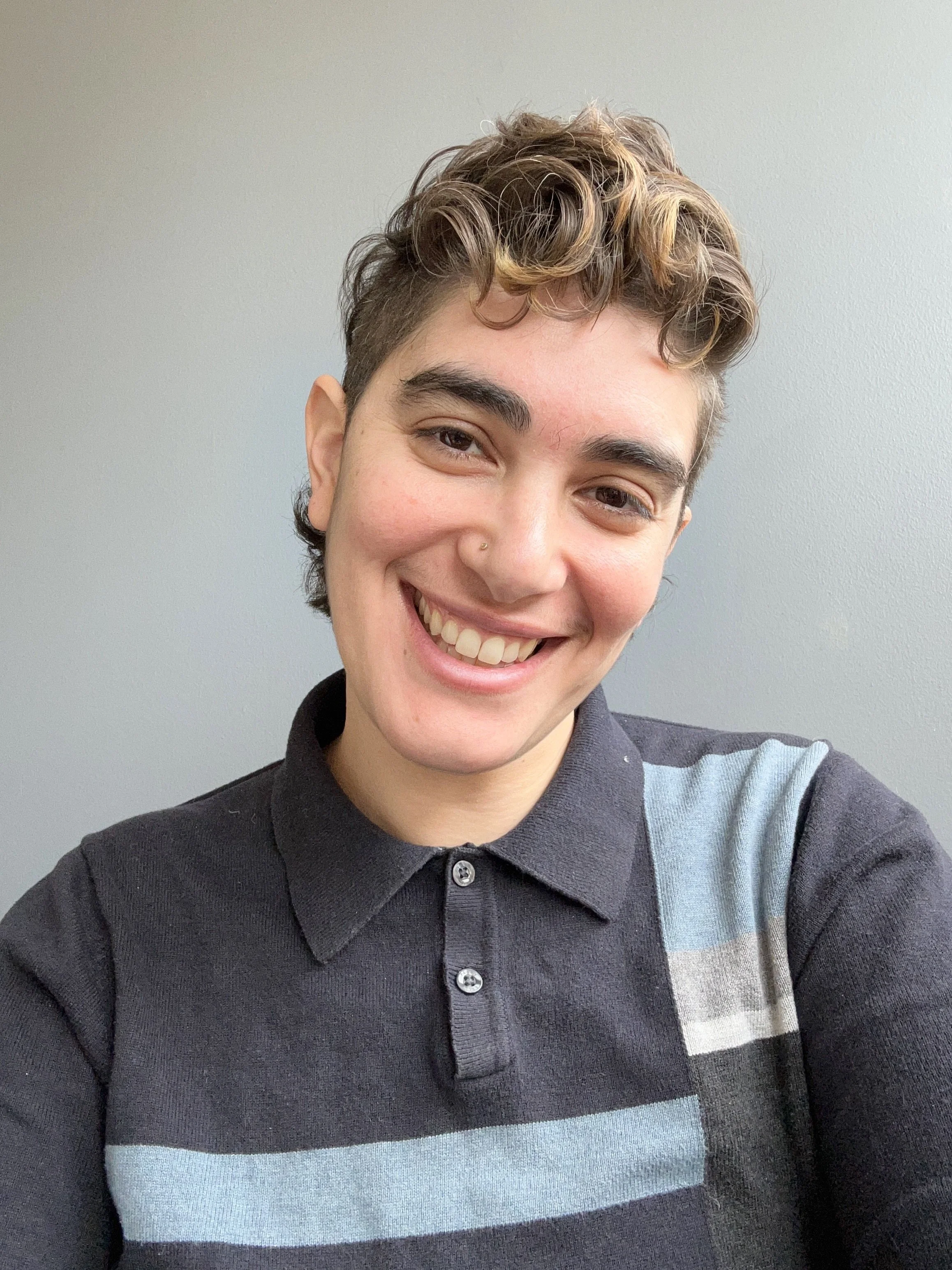 A person with short, curly hair and a nose piercing, smiling and wearing a black collared shirt with light blue and gray stripes, taking a selfie against a plain gray wall.