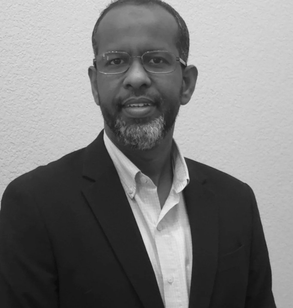 A man with glasses, a beard, and short hair, wearing a suit jacket and a collared shirt, standing in front of a plain wall.