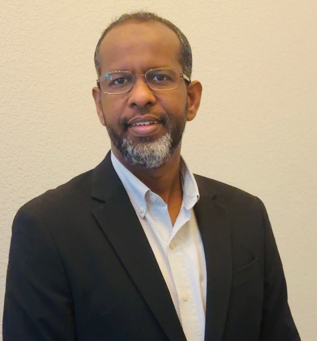 A middle-aged man wearing glasses, a black blazer, and a white shirt, standing against a beige wall.