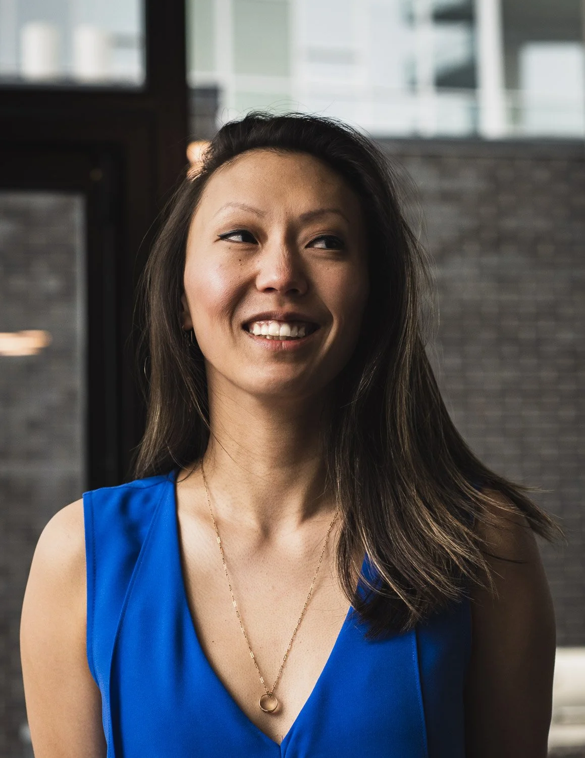 A smiling woman with dark hair wearing a blue sleeveless top and a gold necklace, standing indoors with a window and brick wall in the background.