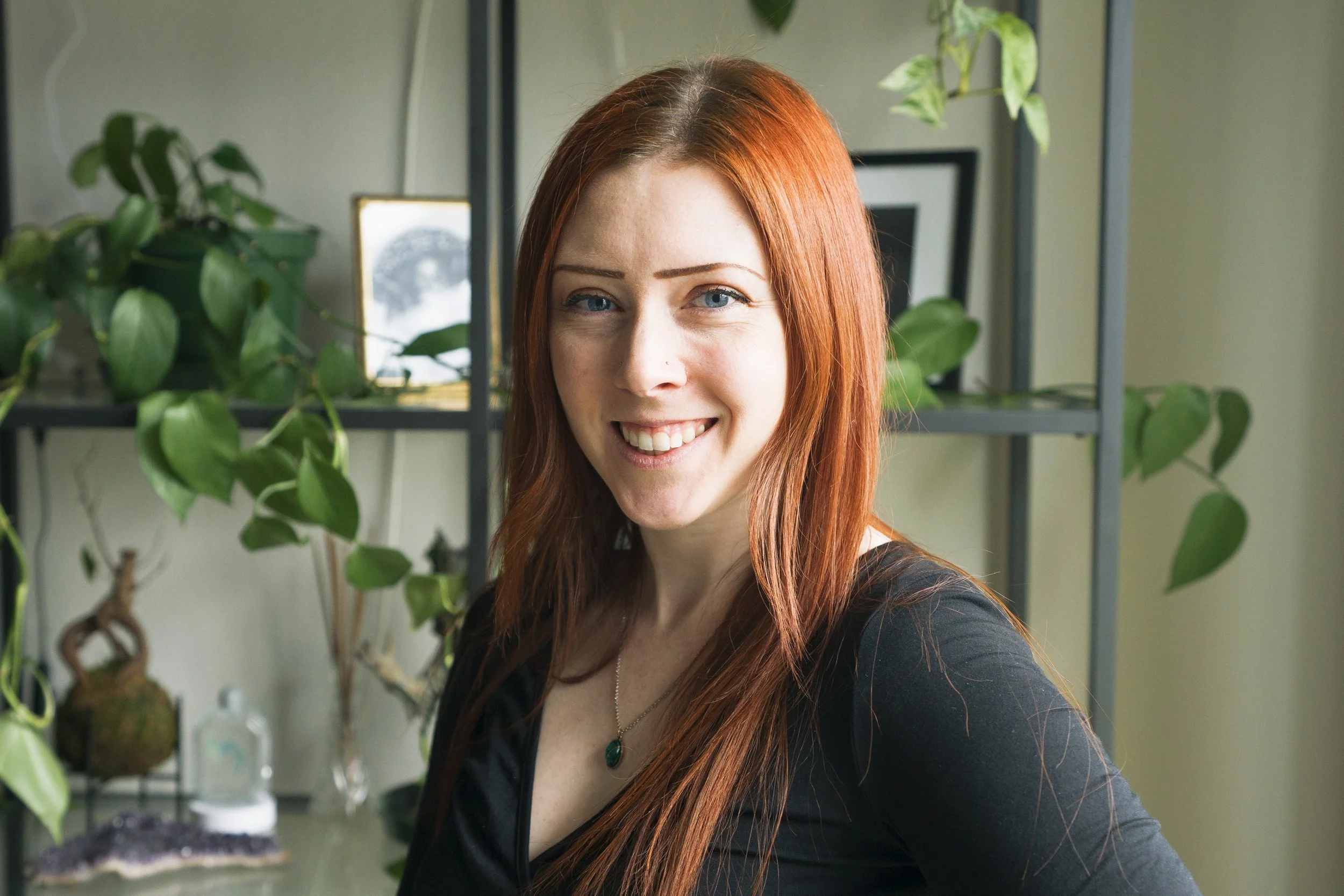 A smiling woman with long red hair and blue eyes, wearing a black top, standing in front of a shelf with green plants and framed pictures.
