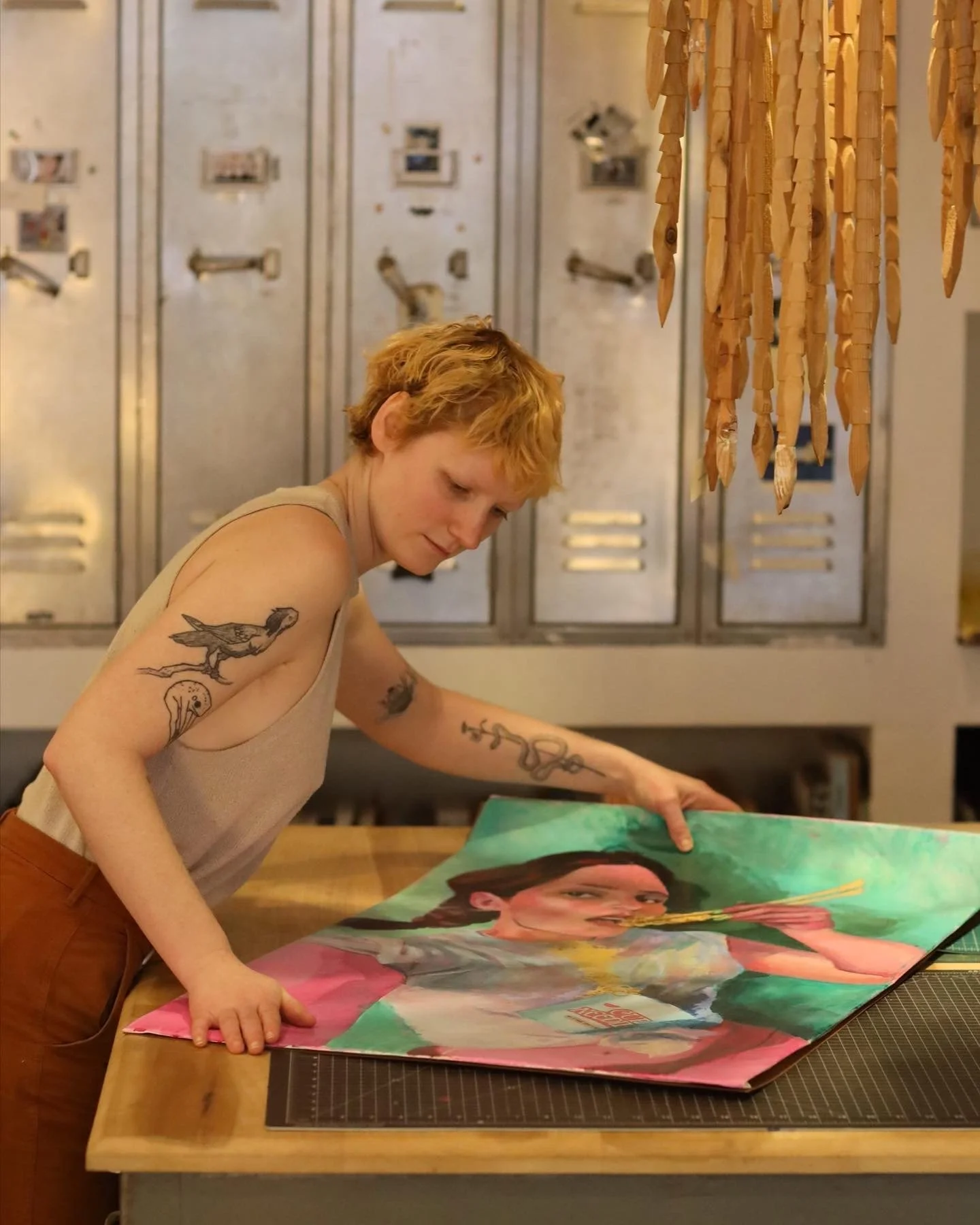 A person with short, curly ginger hair and tattoos on their arms is examining a colorful, portrait-style artwork on a table. The background features gray lockers and hanging wooden art pieces.