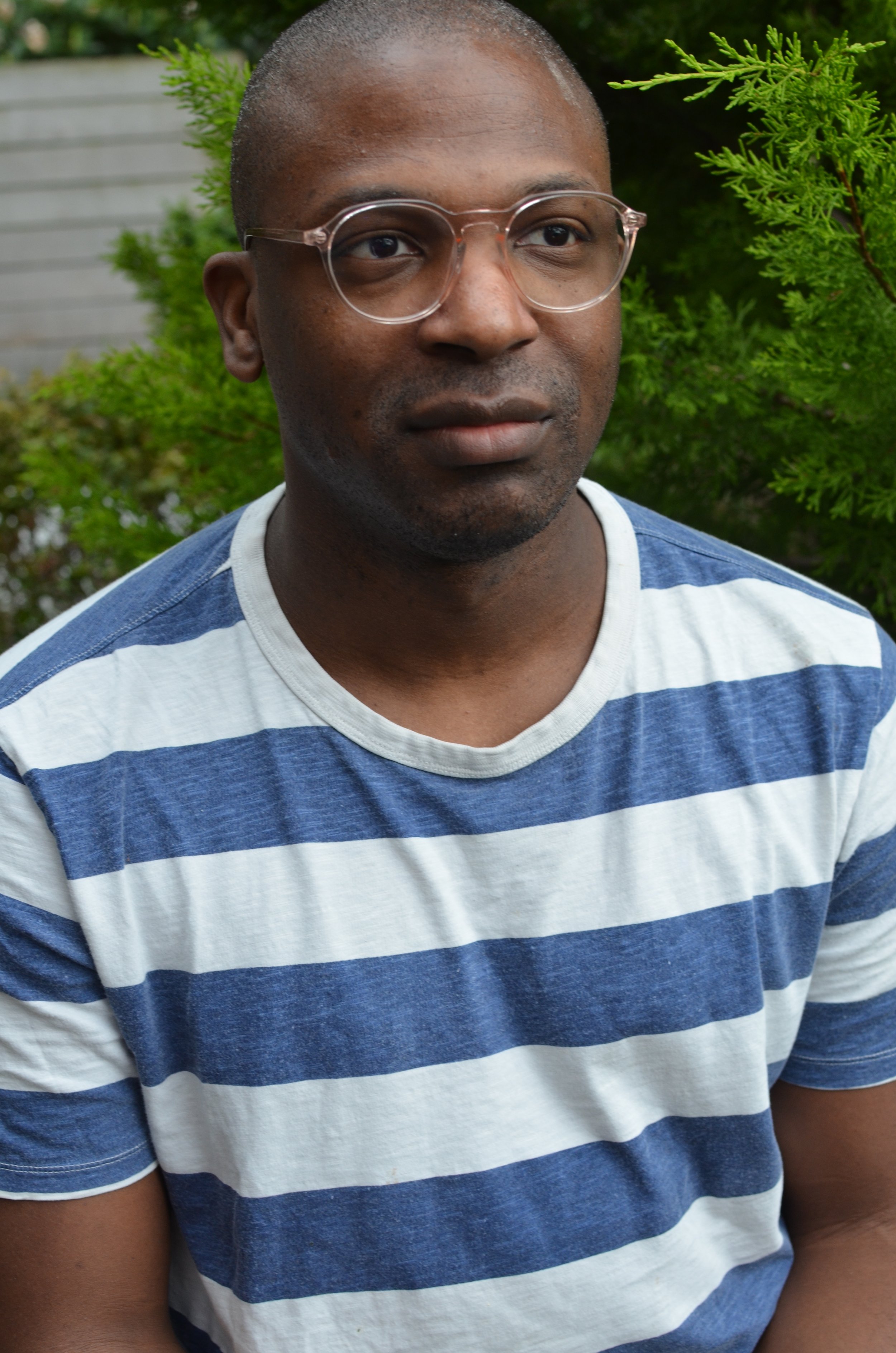 Portrait of a man wearing glasses and a blue and white horizontal striped t-shirt outdoors with green foliage in the background.