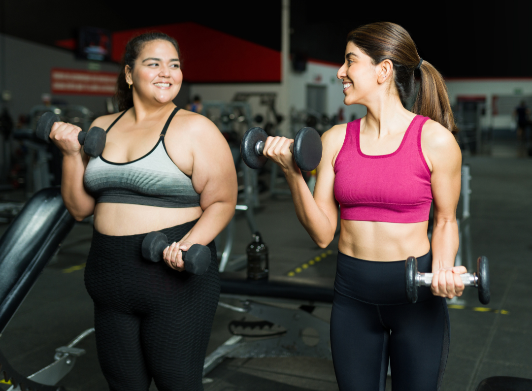 two women in a gym performing biceps curls, both wearing black leggings.  One wearing pink sports bra and one wearing gray sports bra