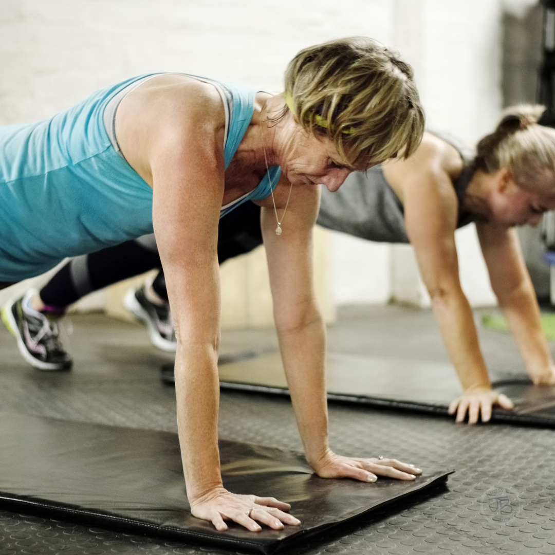 Two women performing a plank exercise on exercise mats during a strength training workout in a gym.