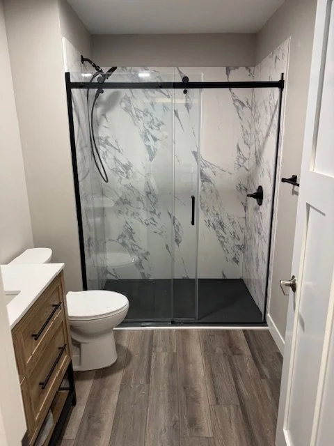 Bathroom with a walk-in shower with glass doors, white marble walls, a toilet, and a wooden vanity.