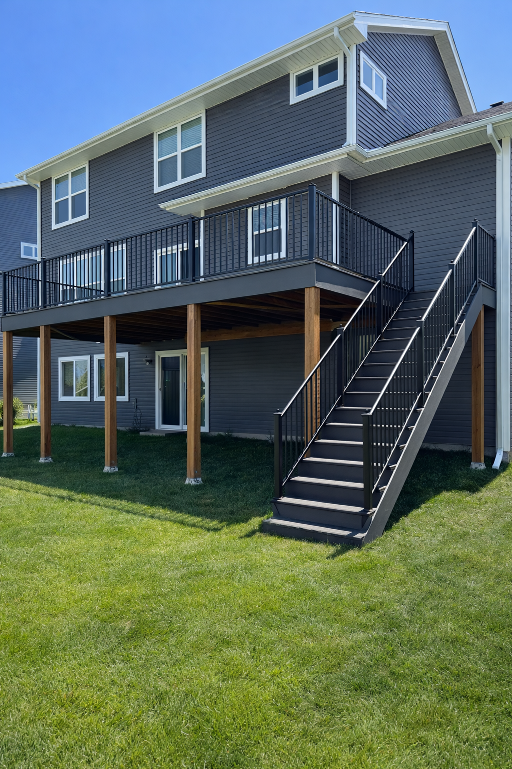 A modern multi-story house with blue siding, multiple white-framed windows, a large upper balcony with black railing, and a staircase leading down to a green grassy yard.
