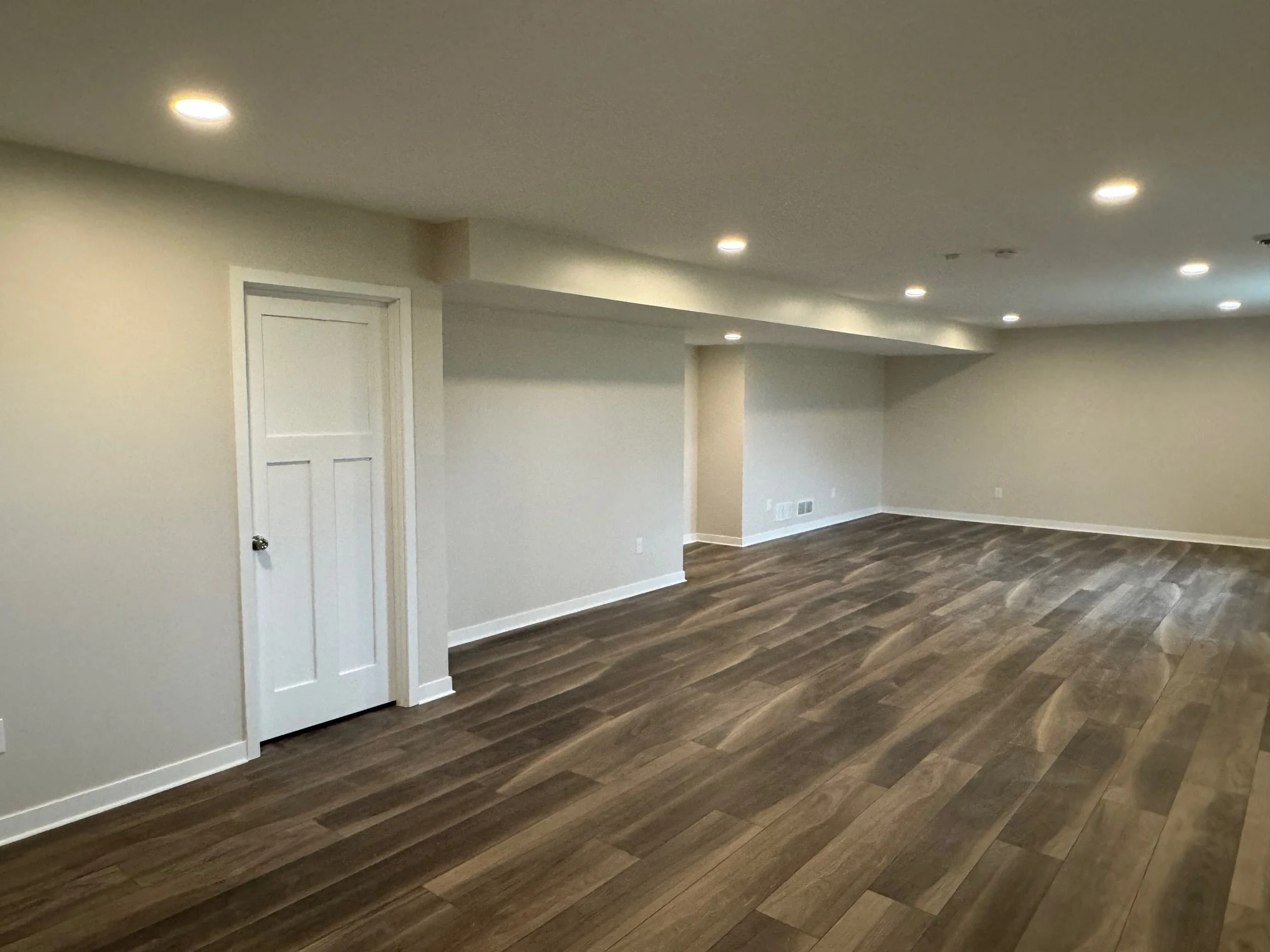 Empty basement room with wood flooring, white walls, and recessed lighting.