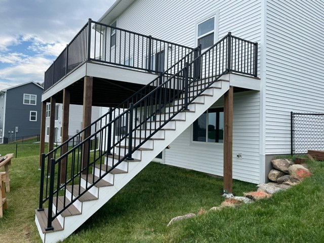 Exterior view of a white house with a composite deck and black metal staircase leading to the upper level, supported by wooden posts, with a grassy yard and neighboring houses visible.