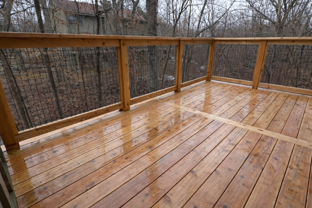 Wooden outdoor deck with a railing, wet from rain, with trees and houses in the background.