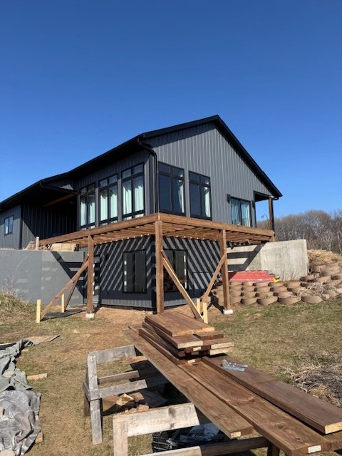 New house under construction with a wooden deck and stairs, set on a hillside with a clear blue sky.
