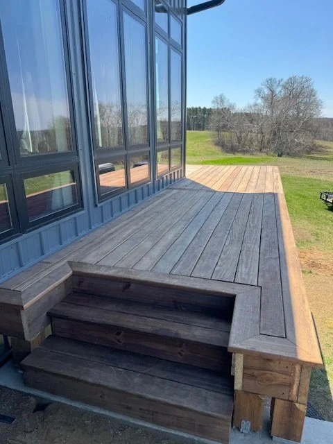 Wooden deck with stairs attached to a house with large windows, overlooking a grassy yard with trees in the background under clear blue sky.