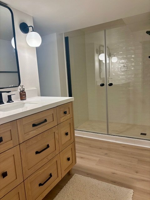 Bathroom with wooden vanity, white countertop, and round white wall sconce light. Glass shower enclosure with sliding doors and white tiled wall.