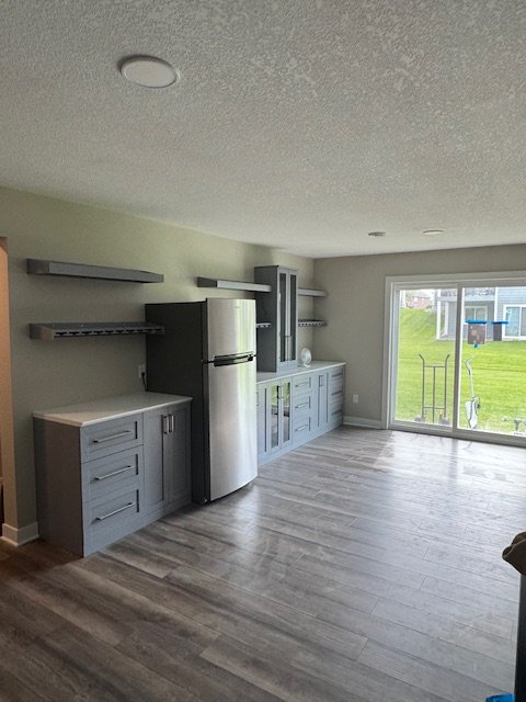 Empty kitchen with gray cabinets, stainless steel refrigerator, open shelves, and sliding glass door overlooking a yard.