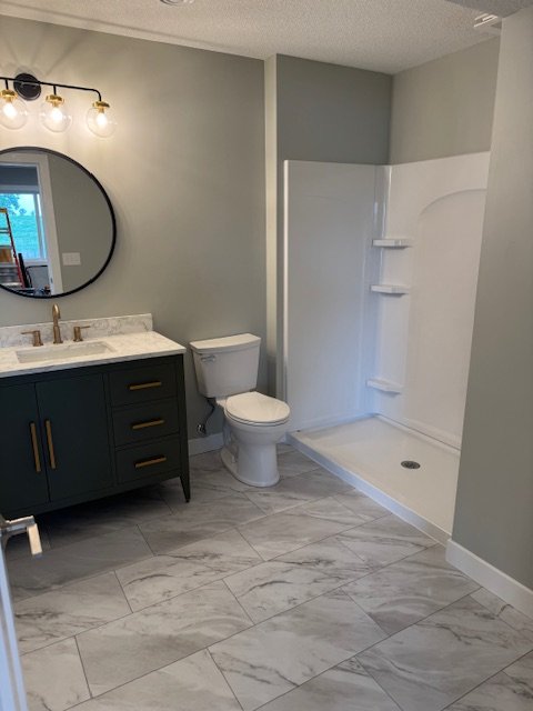 Modern bathroom with a vanity, round mirror, toilet, and a shower area with built-in shelves.