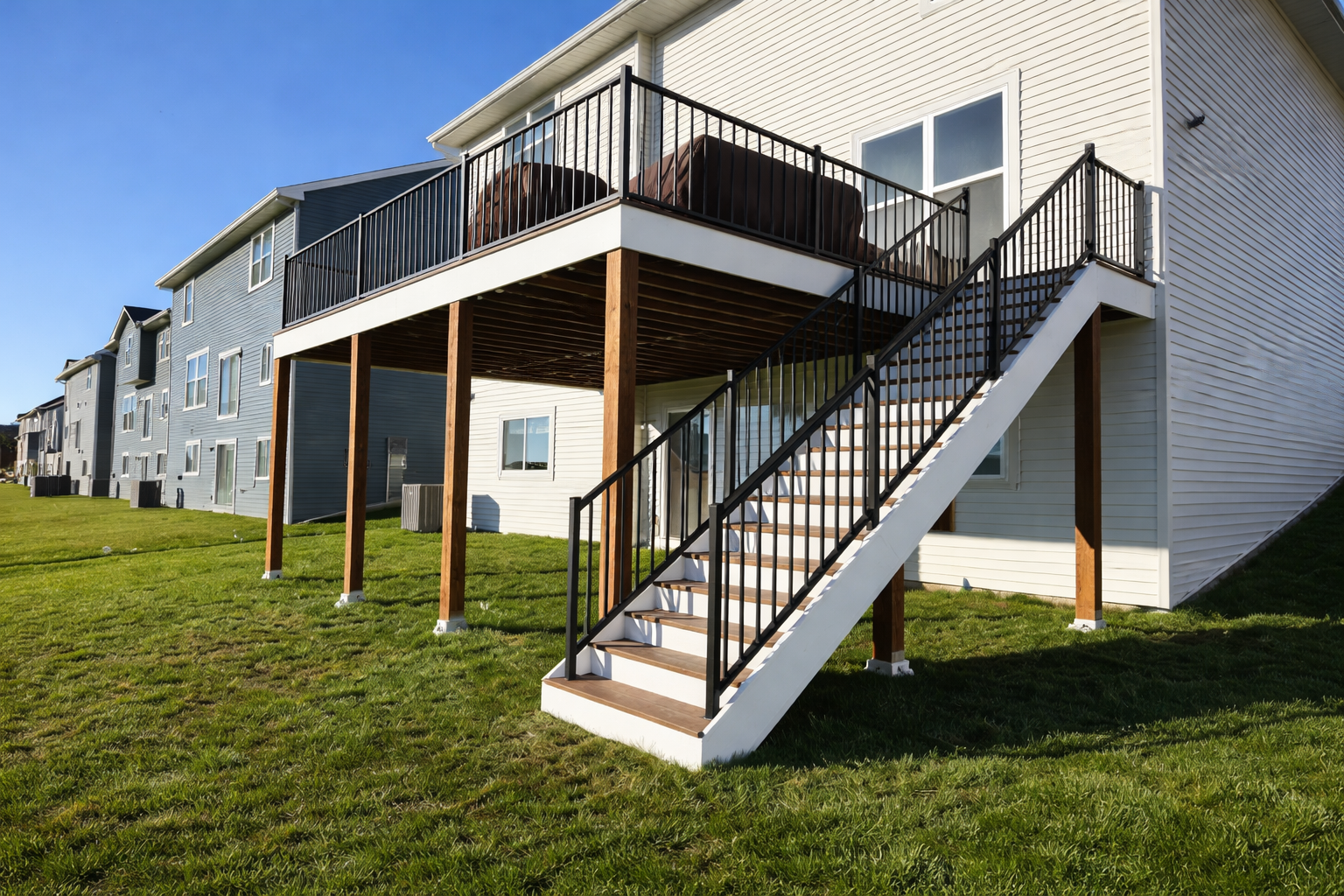 Exterior view of a multi-story residential building with a rear balcony and stairs leading to the ground, surrounded by a grassy yard under a clear blue sky.