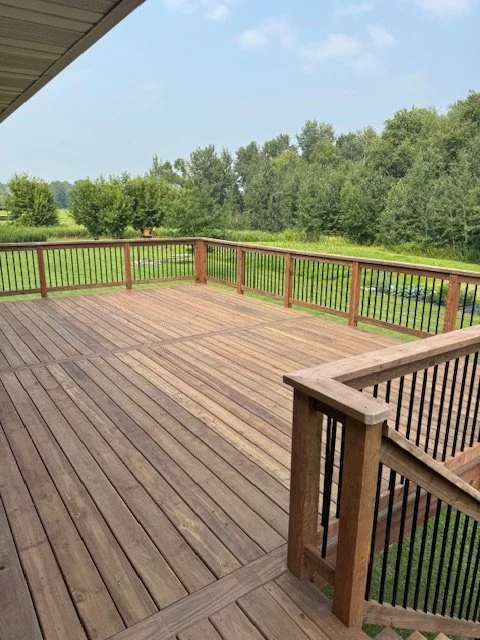 Wooden outdoor deck with railing overlooking a green landscape with trees and blue sky.