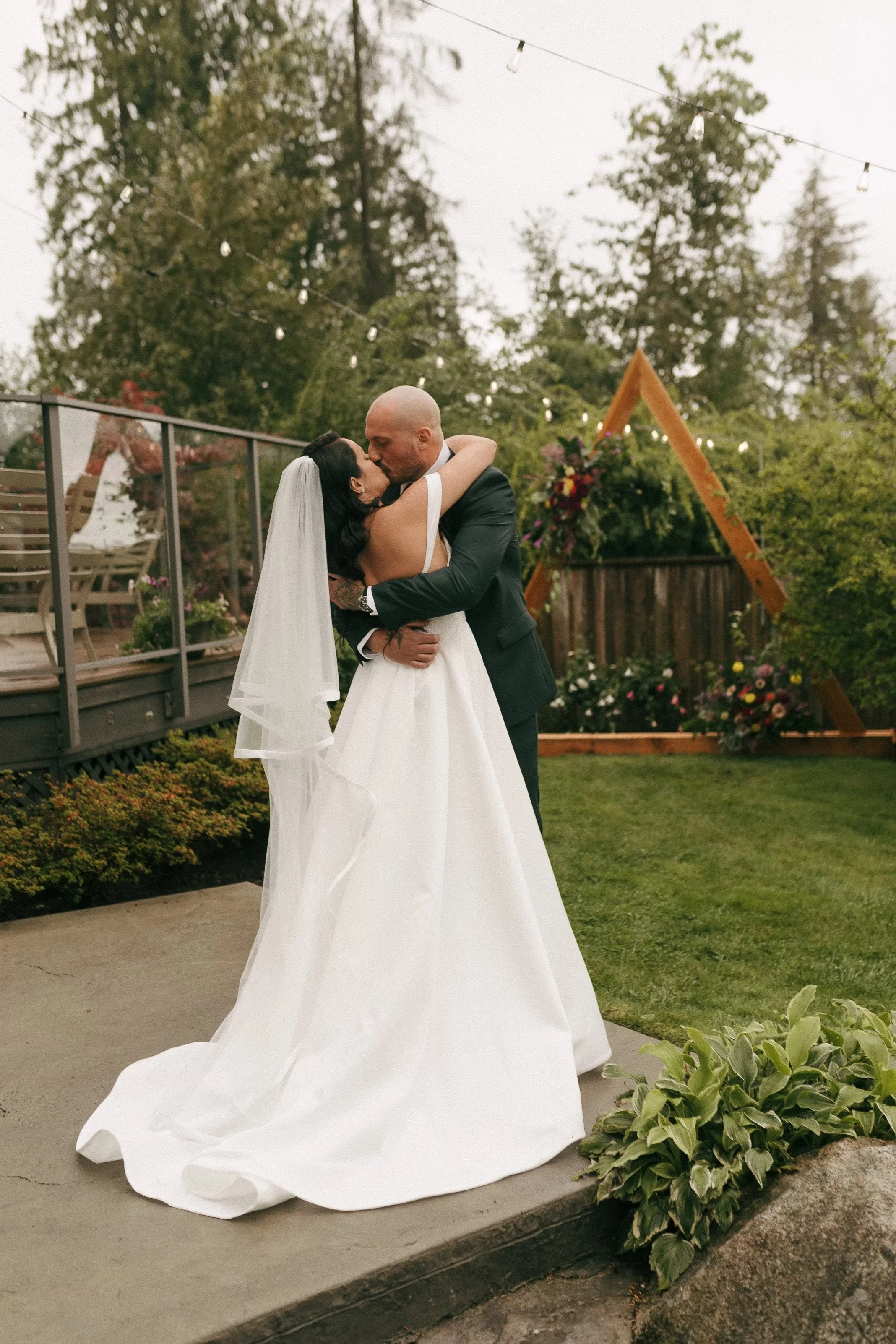 A bride and groom share a kiss during their outdoor wedding ceremony, with string lights and a wooden floral arch in the background, surrounded by trees and greenery.