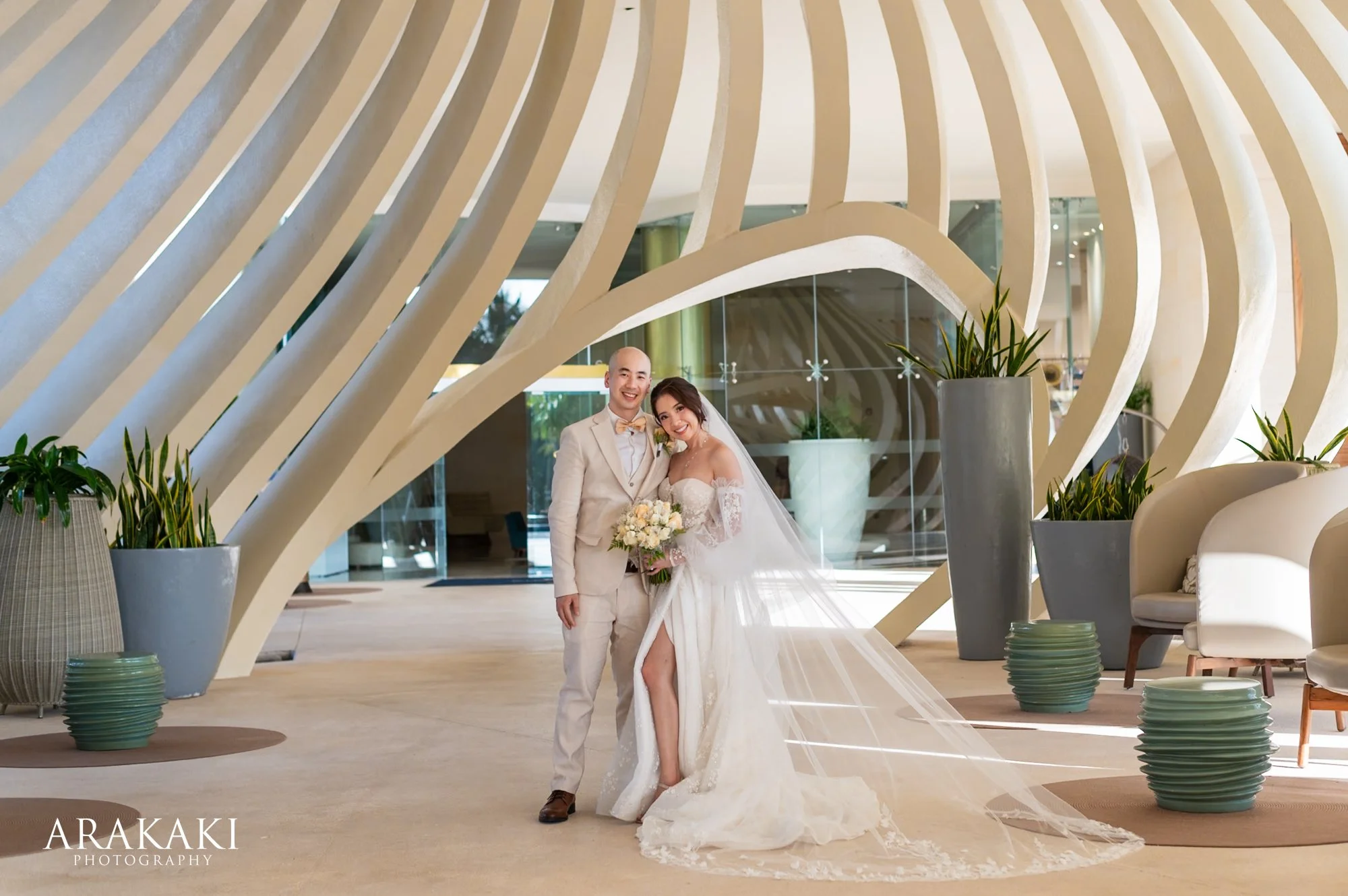 A bride and groom standing together, smiling, inside a modern building with curved architectural features, large planters, and contemporary furniture, during their wedding celebration.