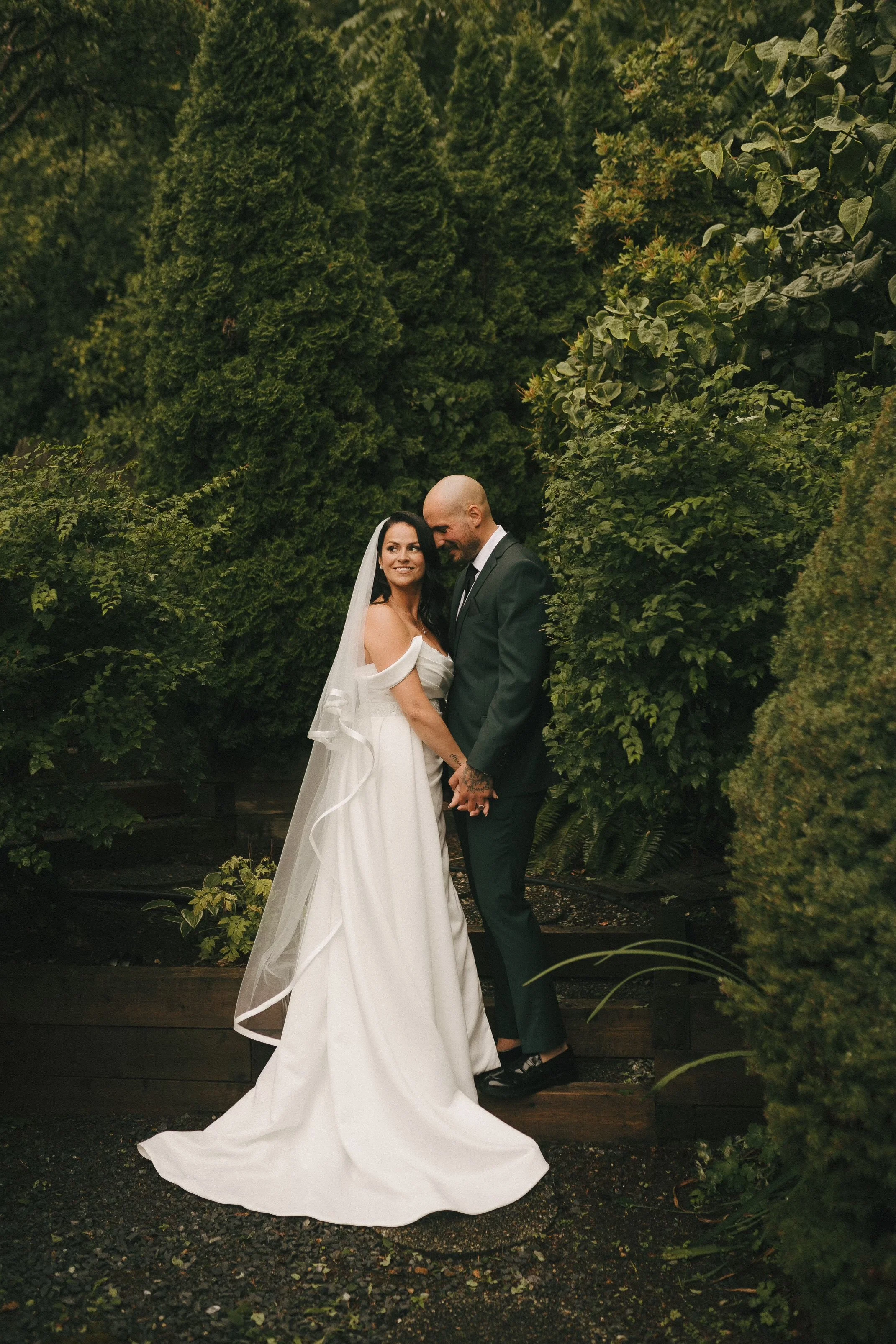 A newlywed couple standing close together outdoors in front of lush green bushes, holding hands and smiling, with the bride in a white off-shoulder wedding gown and veil, and the groom in a black suit with a white shirt and black tie.