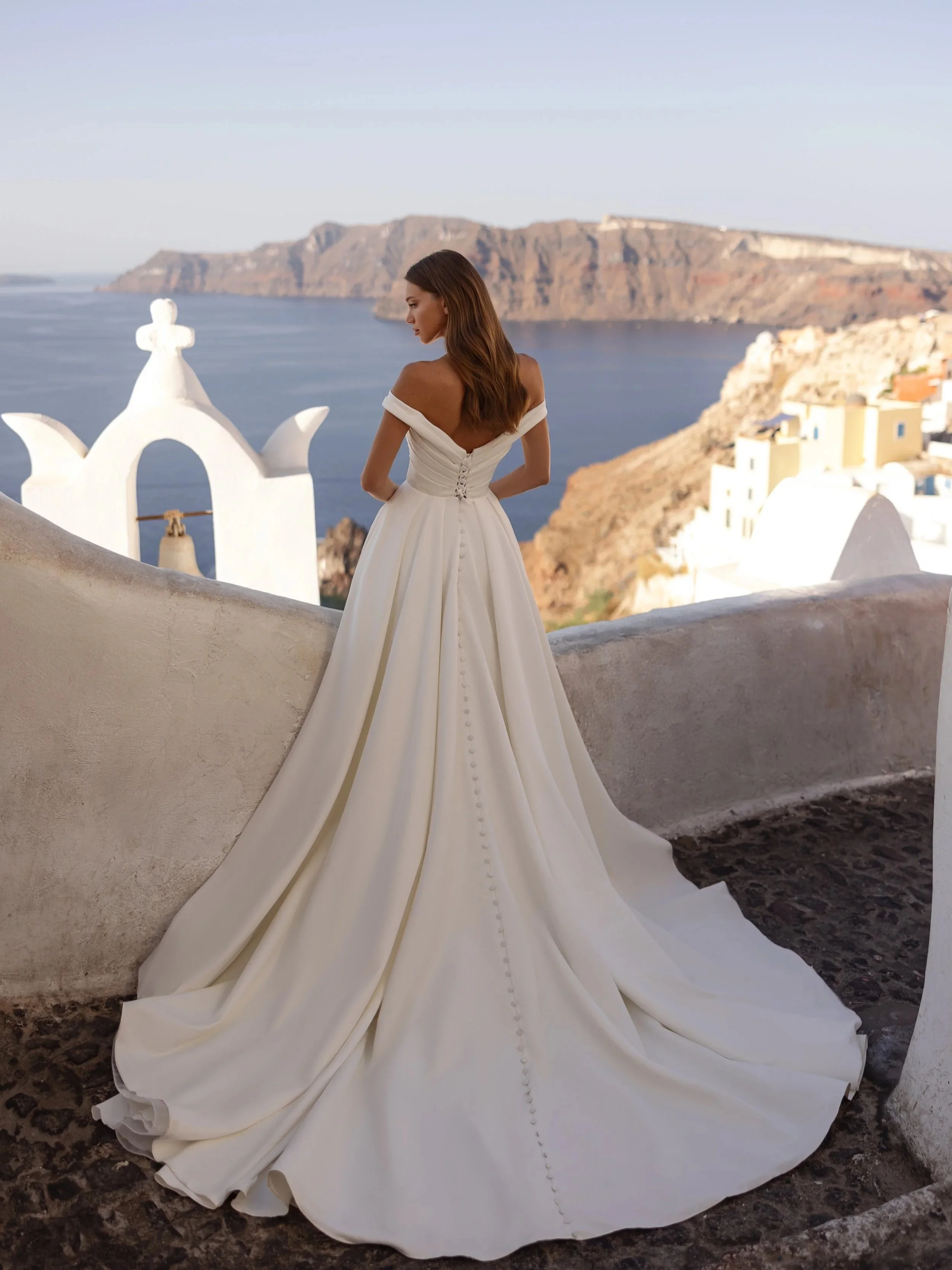 Bride in a Maria Anette off-the-shoulder A-line gown with a long train overlooking the sea in Santorini, Ethereal Bridal, Port Moody