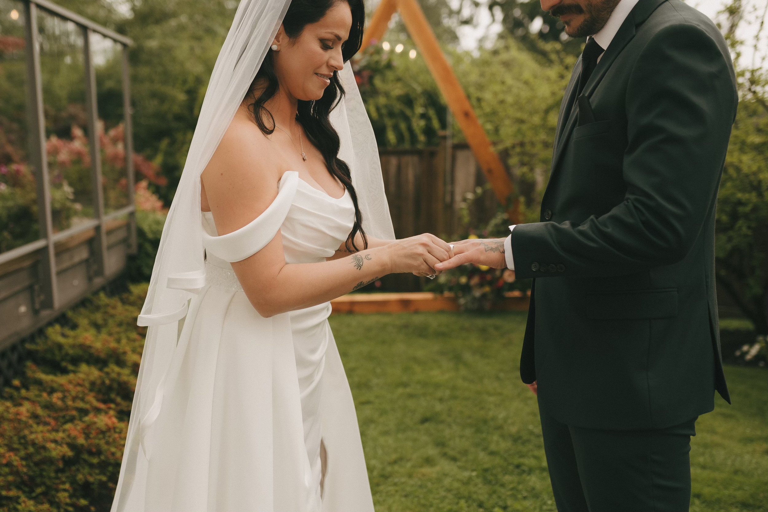 A bride and groom exchanging rings during a wedding ceremony outdoors, with greenery and wooden structures in the background.