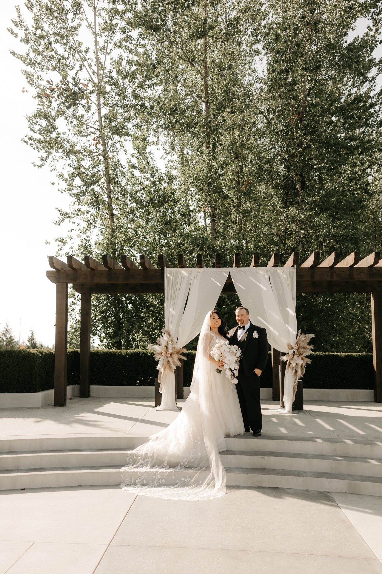 Bride and groom standing together at their outdoor wedding ceremony, with white drapery and floral arrangements behind them, under a wooden pergola with a large tree in the background.
