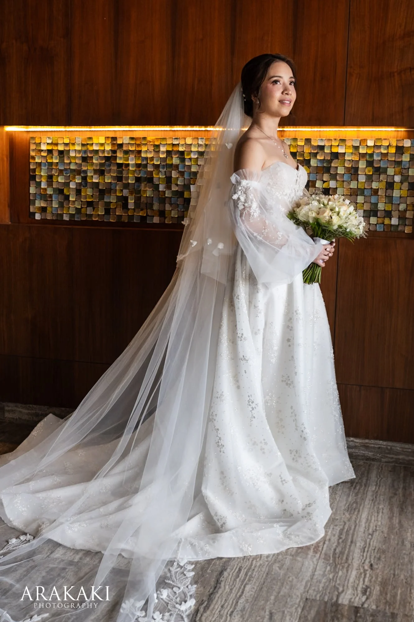 Bride in a white wedding gown with a long veil and bouquet standing indoors against a wooden wall with a decorative panel.