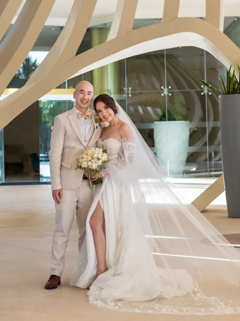 Bride in the 5420 off-the-shoulder lace gown by Maria Anette with a cathedral veil, beside her groom in a modern venue