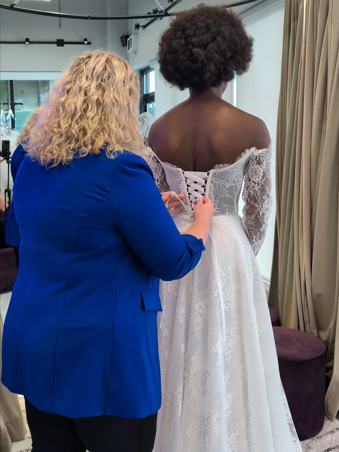 Bridal consultant lacing the corset back of [Gown] by [Designer], an off-shoulder lace A-line wedding dress with long sheer lace sleeves