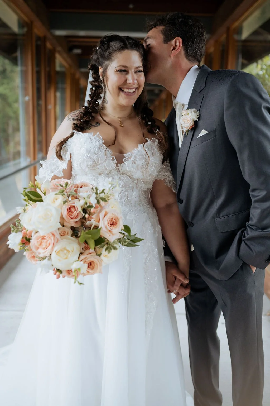 A newlywed couple holding hands, with the groom kissing the bride on her forehead, inside a wooden corridor. The bride is smiling, wearing a white wedding dress with lace details and holding a bouquet of pink and cream roses. The groom is dressed in 