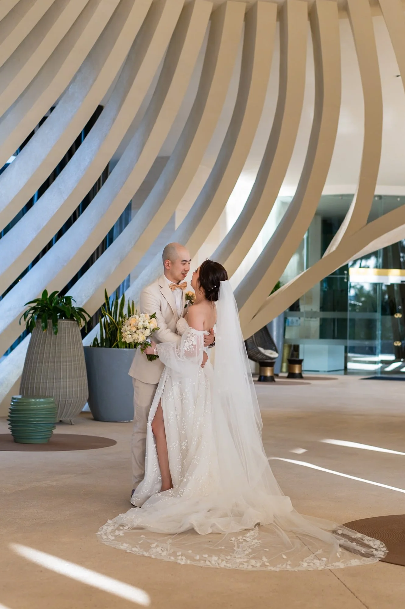 A newlywed couple sharing a moment indoors, with the groom in a beige suit and the bride in a white wedding gown, holding a bouquet of flowers, in front of a modern, wavy architectural background.