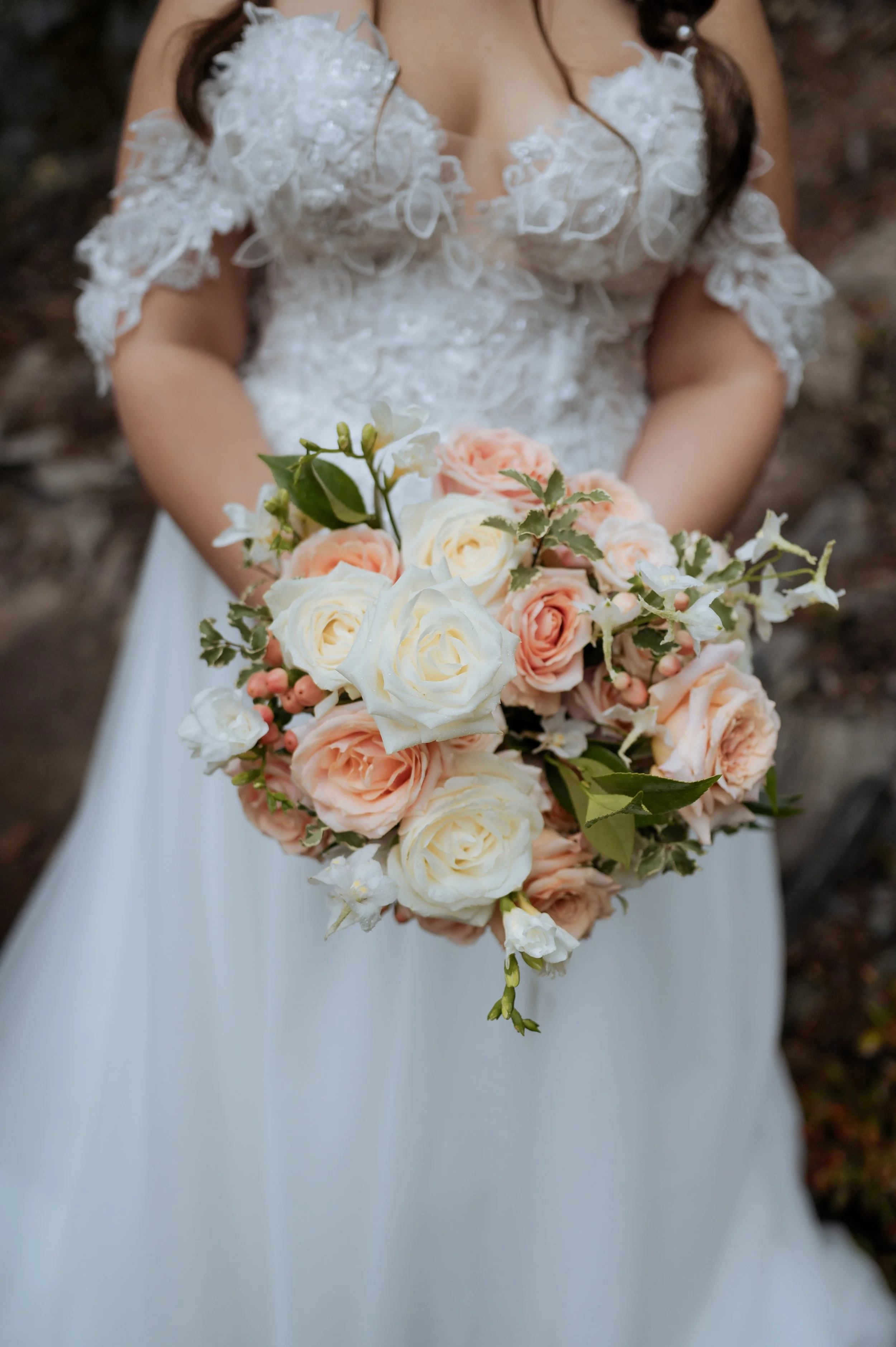 A bride in a white wedding gown with lace details holding a bouquet of white and pale pink roses, white lilies, and greenery.