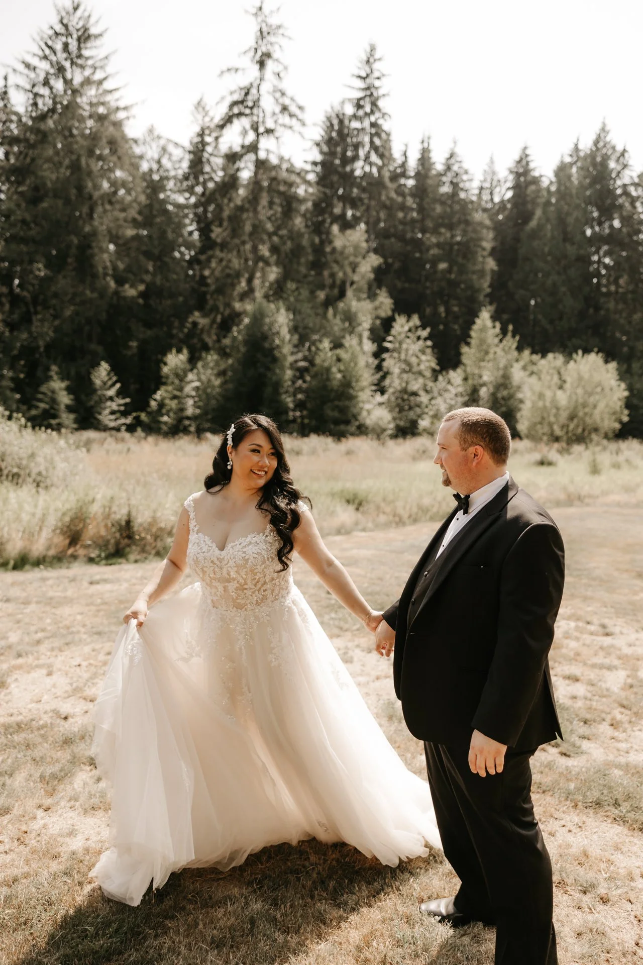 A bride and groom holding hands outdoors on grass, surrounded by trees, smiling at each other on their wedding day.