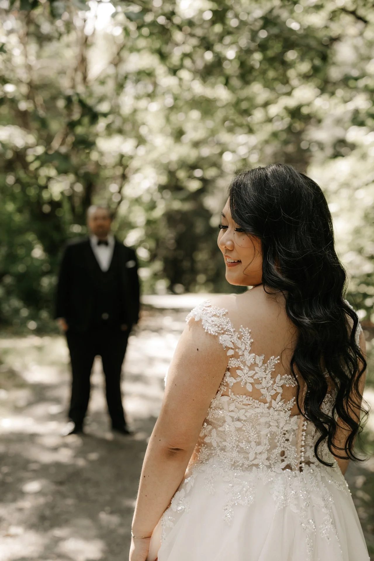 A bride with long black hair wearing a white wedding dress with lace details on her shoulders, smiling and looking down outdoors near a tree, with a man in a black tuxedo standing in the background.