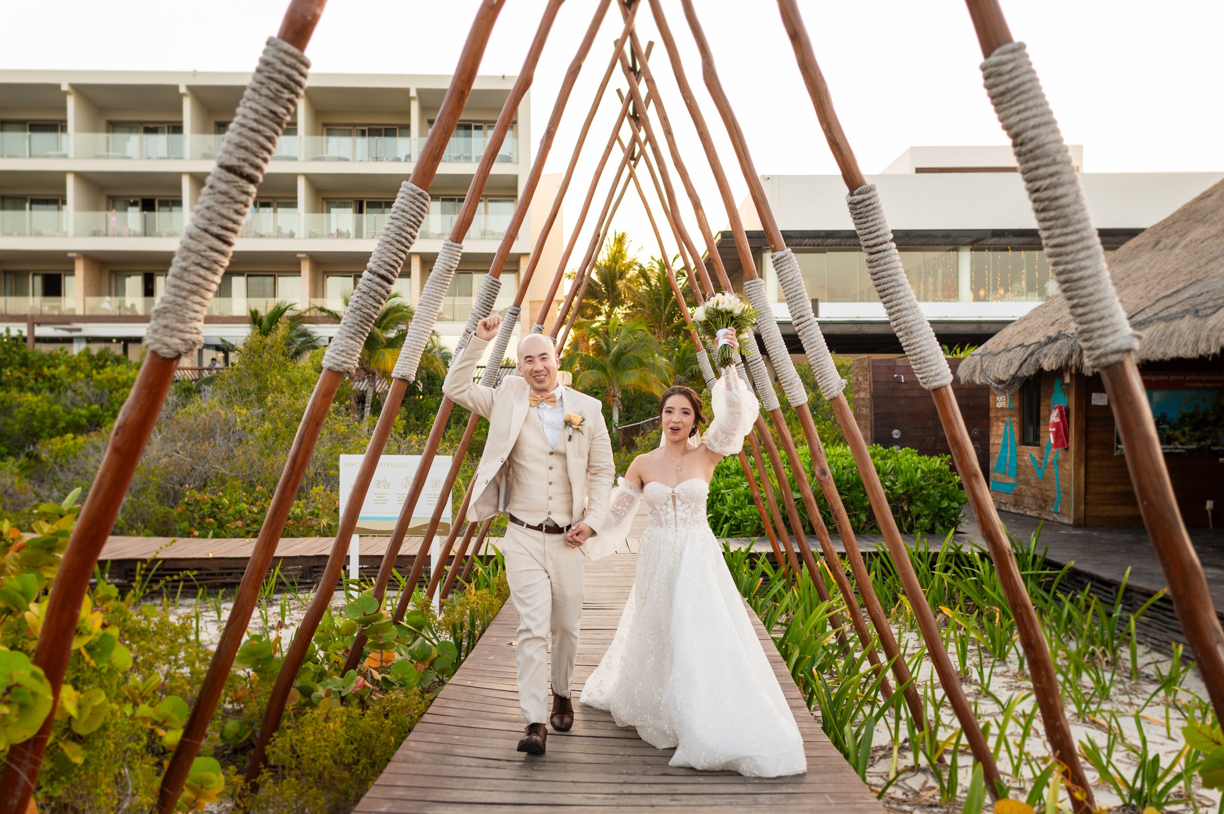 A newlywed couple walking under a teepee structure on a wooden pathway outdoors, holding hands and smiling, with a hotel or resort building and lush green plants in the background.