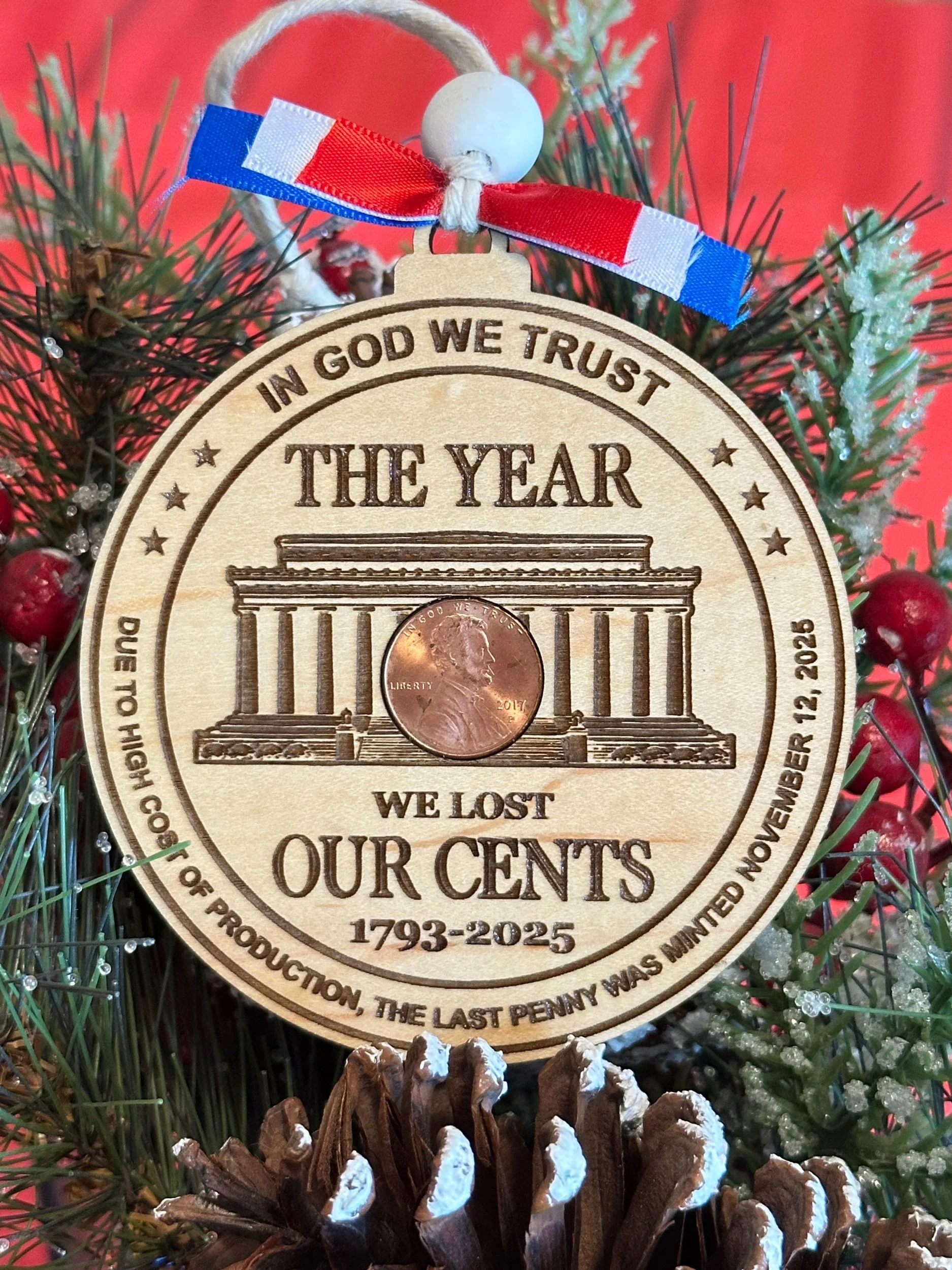 A Christmas ornament shaped like a penny with a penny coin attached, hanging on a decorated Christmas tree. The ornament has an engraved Lincoln Memorial and text referencing the loss of our cents from 1793 to 2025, with a red, white, and blue ribbon.