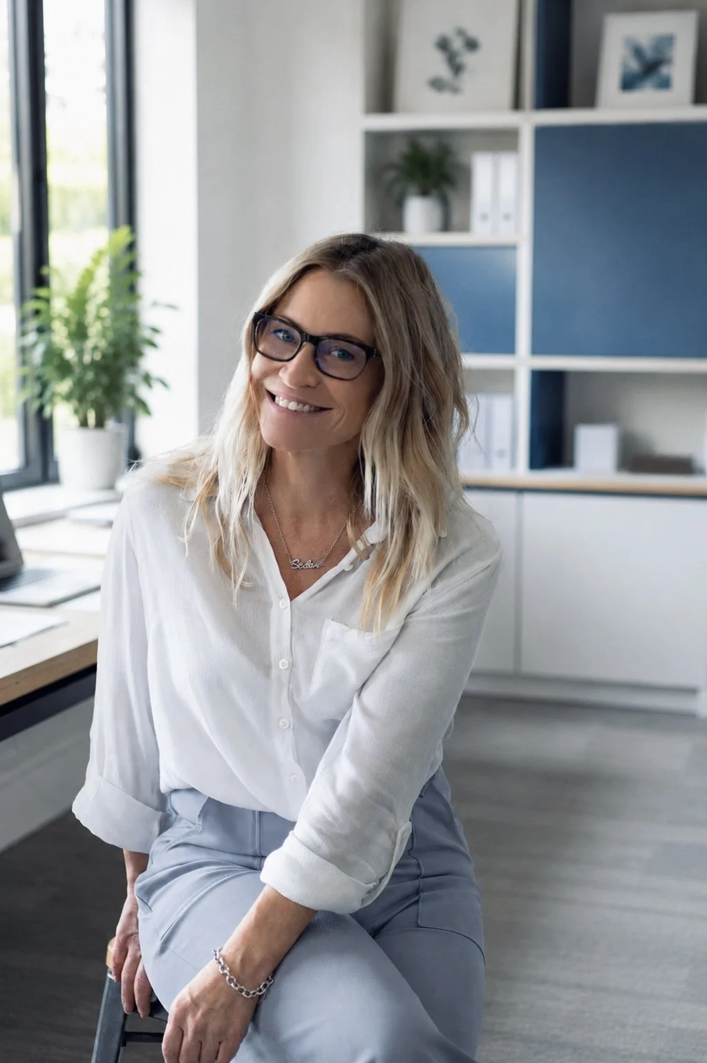 A woman with shoulder-length blonde wavy hair, wearing glasses and a white button-up shirt, sitting on a stool in a modern office space with large windows, plants, and a white bookshelf.